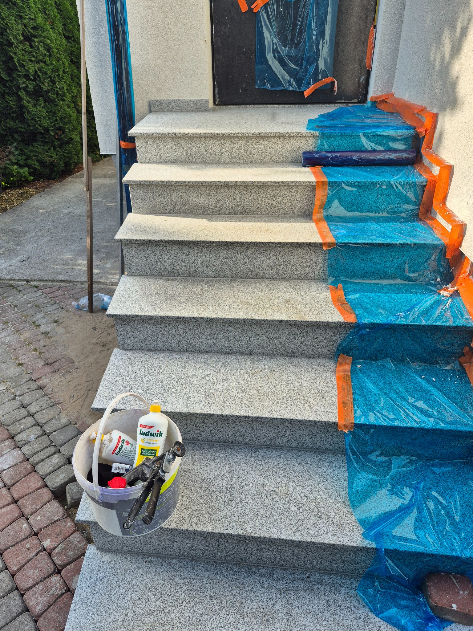 Granitowe schody wejściowe zabezpieczone folią i taśmą malarską podczas prac. Wiadro z narzędziami i preparatami czyszczącymi obok schodów.