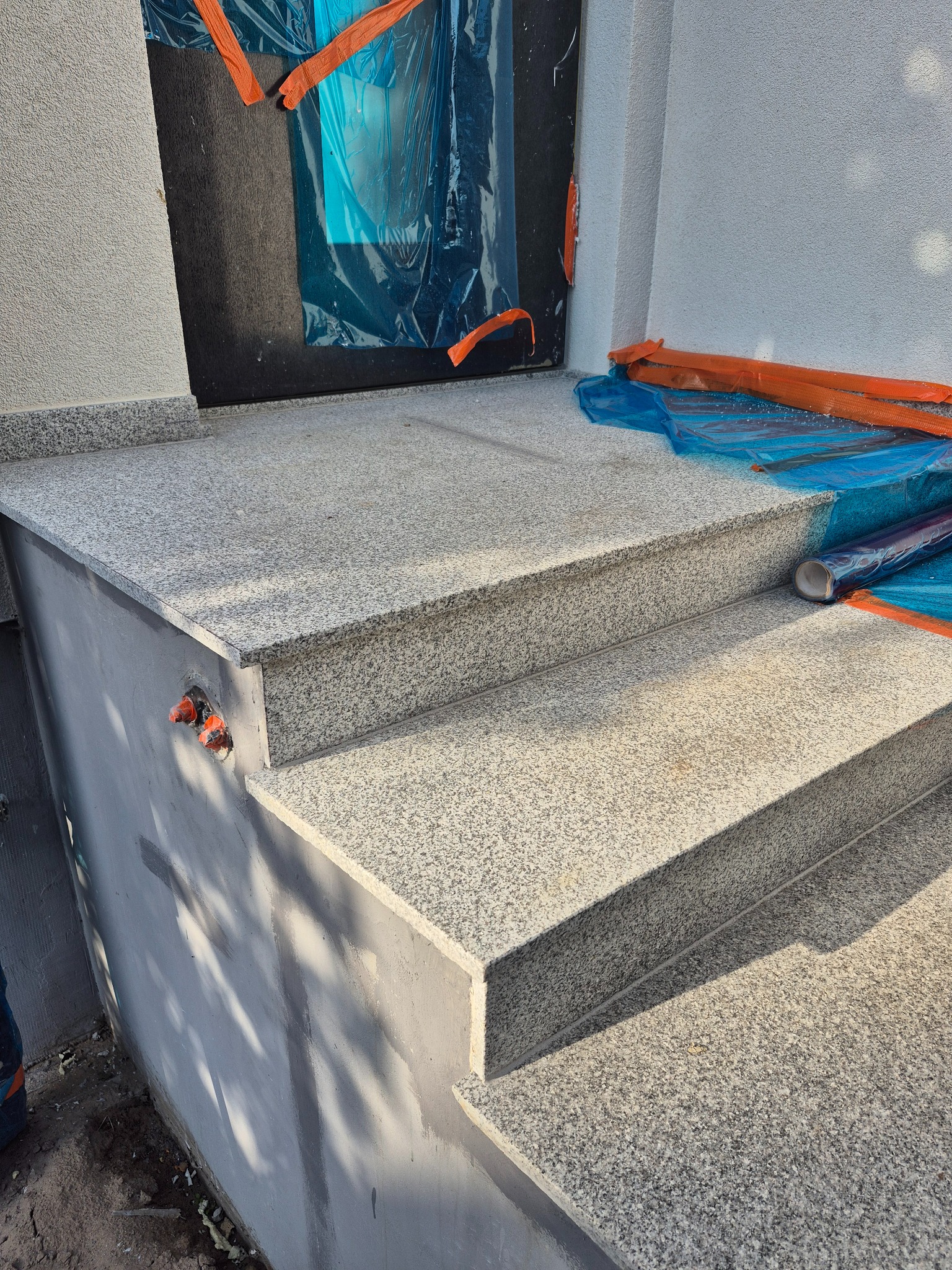 Granitowe schody wejściowe, zabezpieczone folią ochronną, z widocznymi elementami instalacji elektrycznej na ścianie. Jasna, ziarnista faktura kamienia, delikatne cieniowanie.