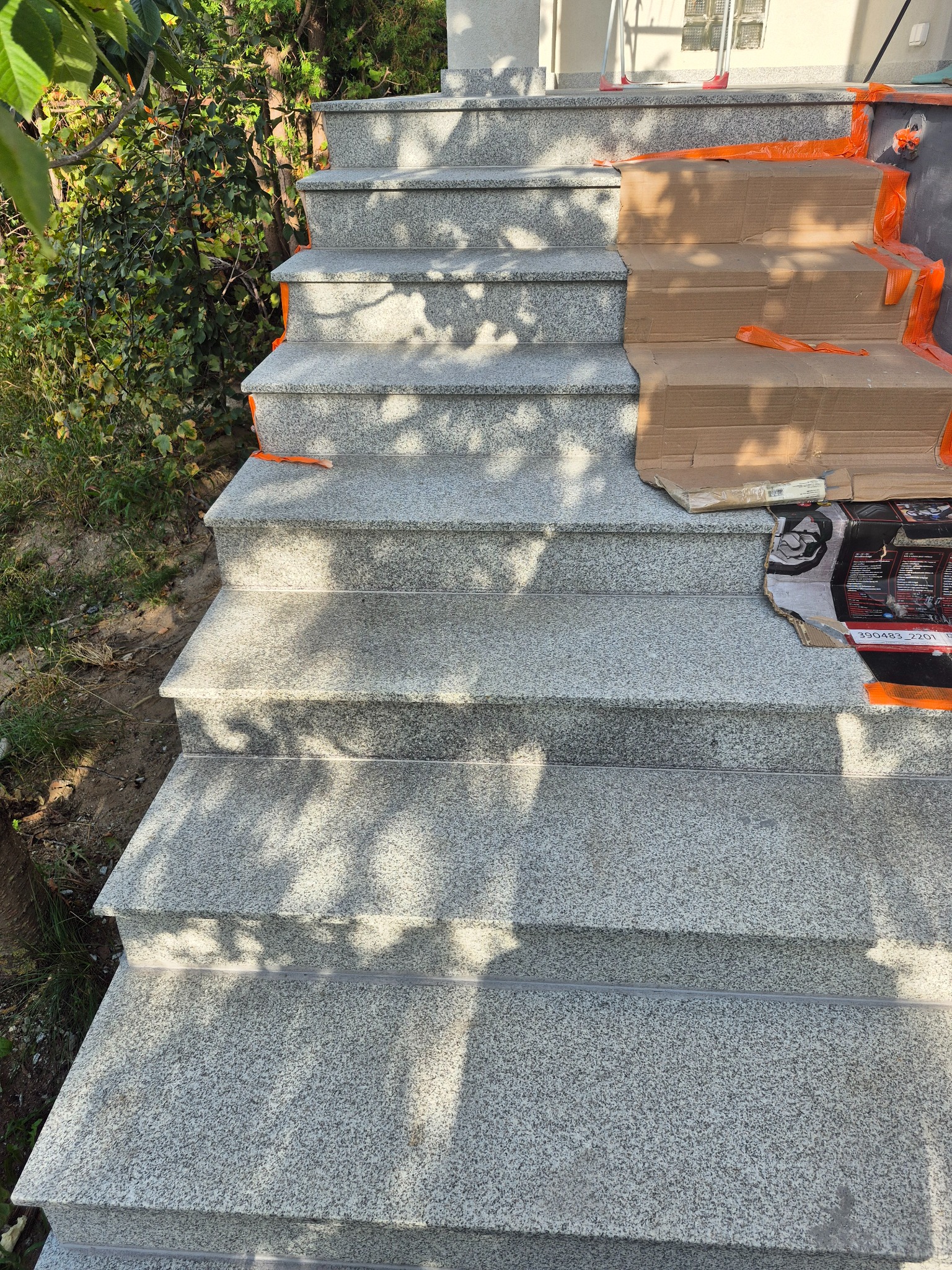 Granitowe schody zewnętrzne w trakcie montażu, częściowo zabezpieczone kartonem i pomarańczową taśmą. Widoczne cienie drzew.