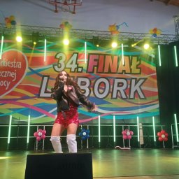 Marta Głodowska -Piosenkarka - Pop-Rock - Wokalistka w cekinowym stroju na scenie podczas 34. Finału WOŚP w Lęborku, oświetlona reflektorami, z mikrofonem w dłoni, w dynamicznej pozie.