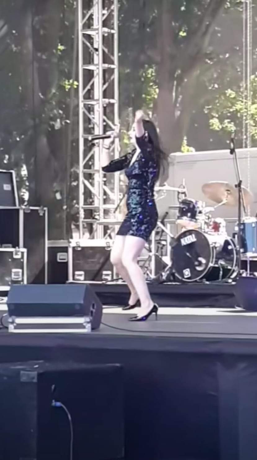 Wokalistka w cekinowej sukience i czarnych szpilkach, dynamicznie śpiewa na scenie podczas koncertu plenerowego, widoczny zestaw perkusyjny Natal.