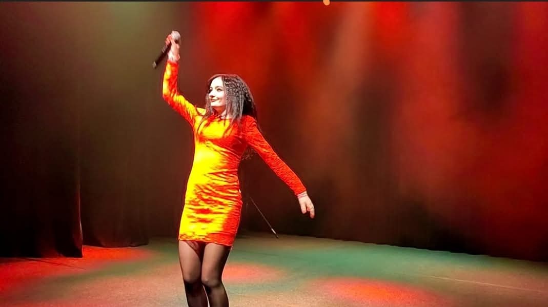 Uśmiechnięta kobieta w pomarańczowej sukience, trzymająca mikrofon na scenie z czerwonym tłem i zieloną podłogą.