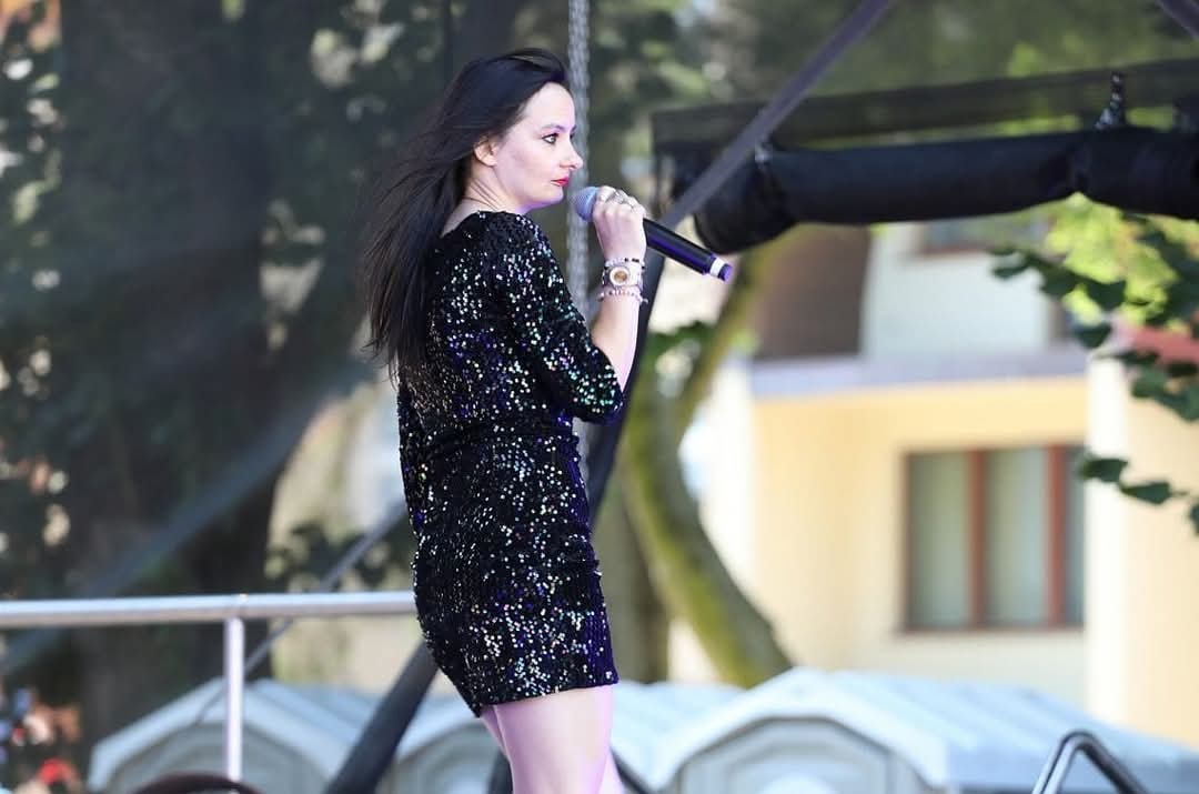 Kobieta w cekinowej sukience śpiewa do mikrofonu na scenie plenerowej, w tle budynek i drzewa.