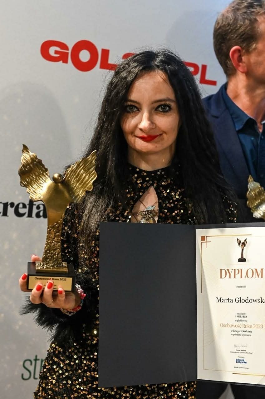 Portret kobiety z ciemnymi włosami, trzymającej złotą statuetkę i dyplom Osobowości Roku 2023 w kategorii Kultura, w błyszczącej sukience na tle logo.