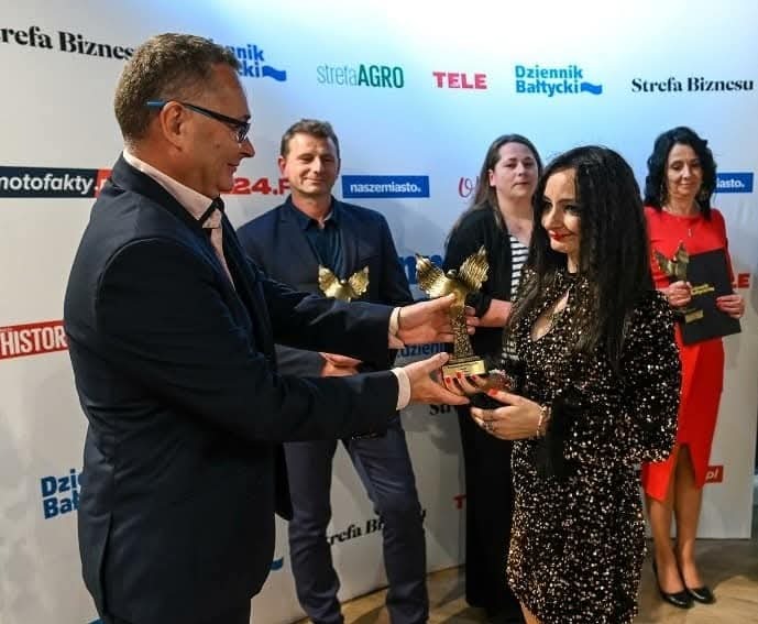 Kobieta w cekinowej sukience odbiera nagrodę w kształcie złotego orła na tle ściany z logo sponsorów.