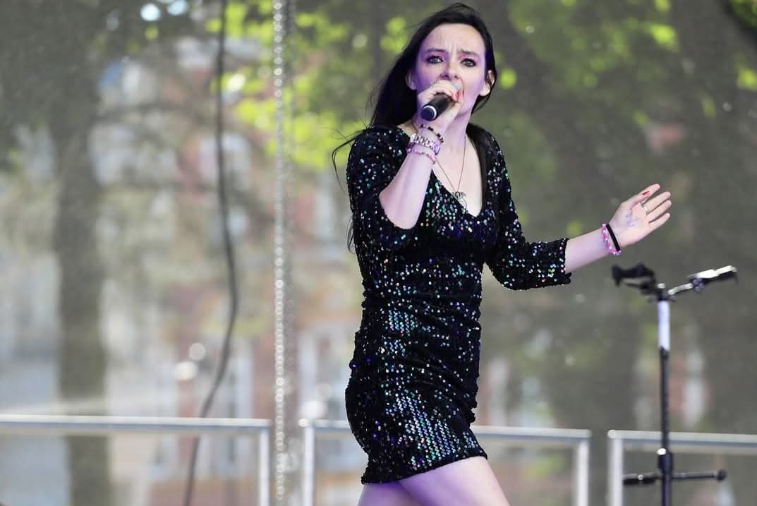 Kobieta w cekinowej sukience śpiewa do mikrofonu na scenie, z widocznym statywem mikrofonowym i rozmytym tłem z zielenią drzew.