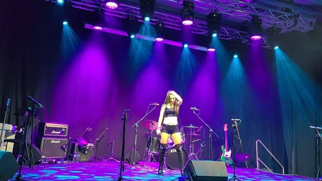 Wokalistka w czarnym stroju scenicznym na scenie oświetlonej niebiesko-fioletowymi światłami, z widocznymi instrumentami i wzmacniaczami Marshall.