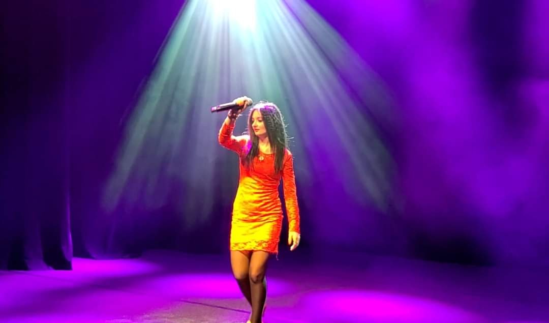 Wokalistka w pomarańczowej sukience śpiewa na scenie oświetlonej fioletowymi i białymi reflektorami, trzymając mikrofon w uniesionej dłoni.