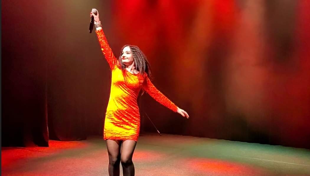 Kobieta w pomarańczowej sukience i czarnych rajstopach śpiewa na scenie, trzymając mikrofon w uniesionej ręce, oświetlona czerwonym światłem.