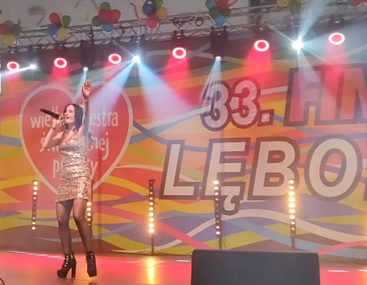 Wokalistka w złotej sukience śpiewa na scenie podczas koncertu WOŚP Lebork, z mikrofonem w ręku i podniesioną dłonią.