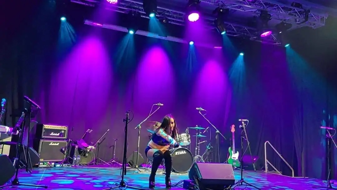 Wokalistka na scenie w blasku fioletowych świateł, otoczona instrumentami i mikrofonami. W tle perkusja i kurtyna.