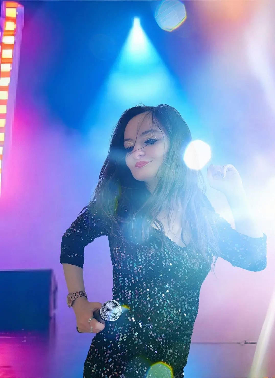 Uśmiechnięta kobieta w cekinowej sukience śpiewa do mikrofonu na scenie w niebieskim oświetleniu. Widoczny zegarek na ręce i delikatny makijaż.