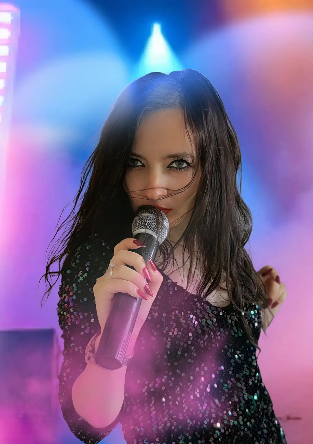 Ujęcie wokalistki z mikrofonem na tle sceny z efektami świetlnymi. Ciemne włosy, błyszcząca sukienka, czerwone usta. Koncert w blasku reflektorów.