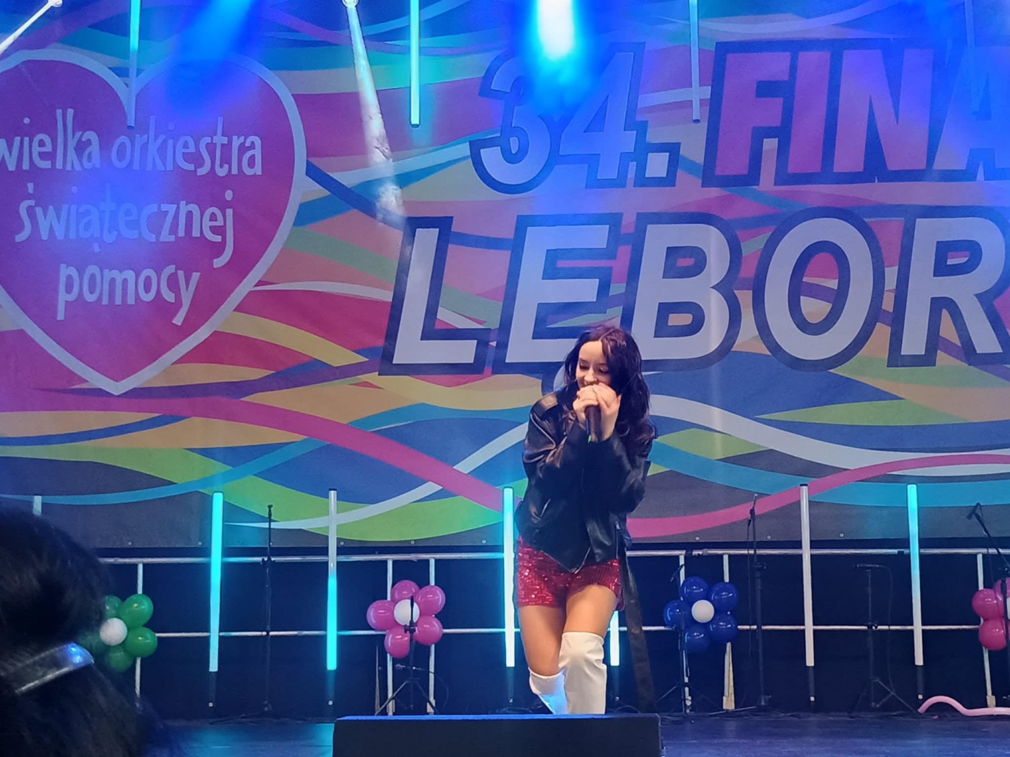 Piosenkarka na scenie podczas 34. Finału WOŚP Lebork, w skórzanej kurtce i cekinowych szortach, śpiewa do mikrofonu na tle dekoracji i balonów.