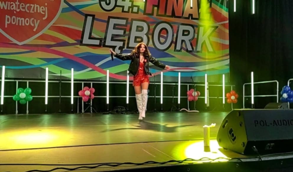 Wokalistka w czerwonej sukience i białych kozakach na scenie podczas koncertu WOŚP w Łeborku. W tle dekoracje i sprzęt nagłośnieniowy.