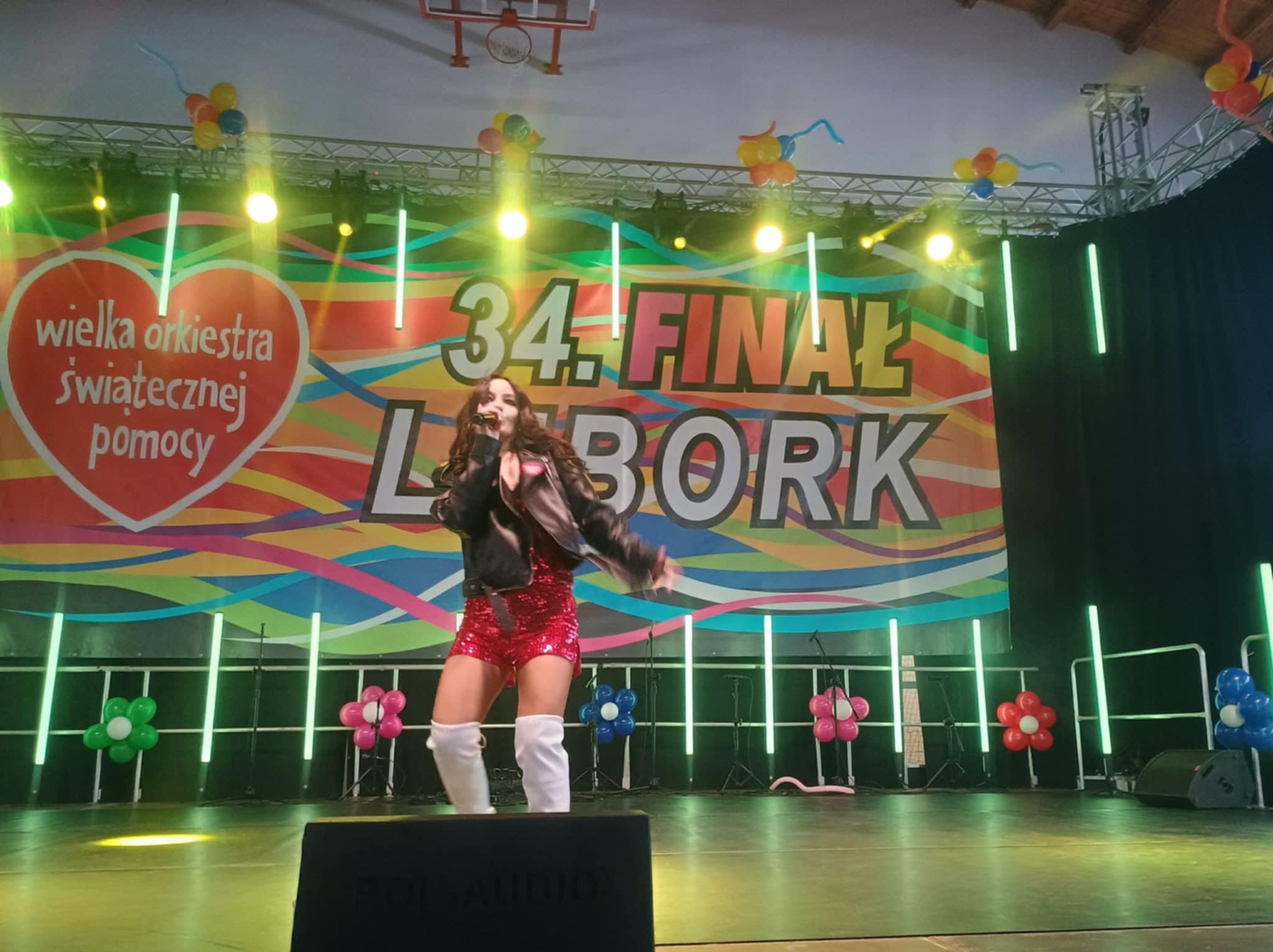 Wokalistka w cekinowym stroju na scenie podczas 34. Finału WOŚP w Lęborku, oświetlona reflektorami, z mikrofonem w dłoni, w dynamicznej pozie.