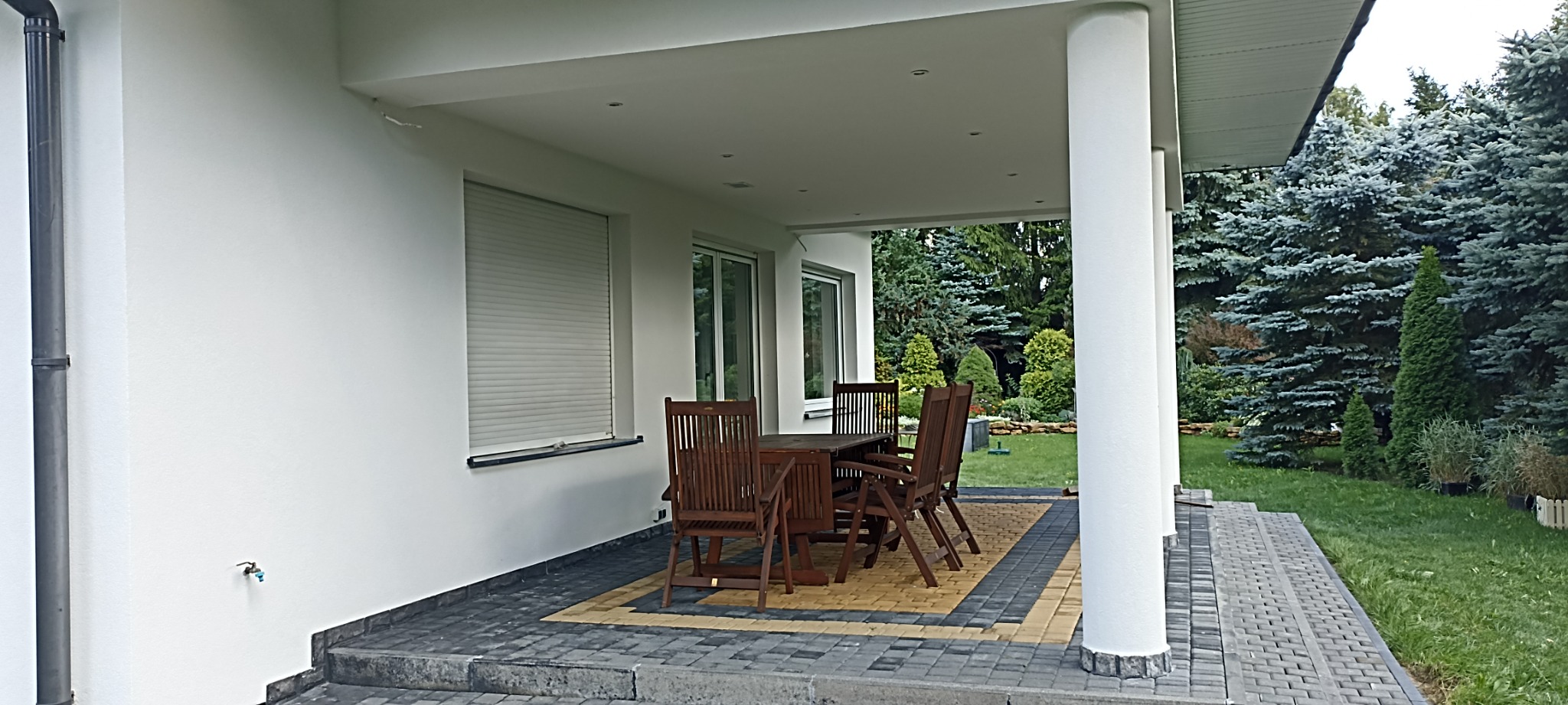 Zadaszony taras z drewnianymi meblami ogrodowymi, białe ściany budynku z roletami, widok na zadbany ogród z iglakami w tle.