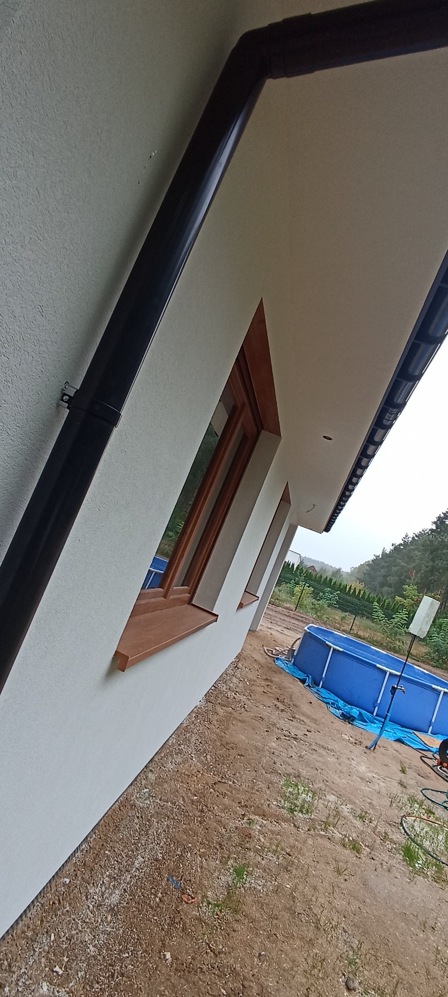 Ściana budynku po ociepleniu z widocznymi oknami z drewnianymi ramami, rynnami i fragmentem basenu ogrodowego w tle.