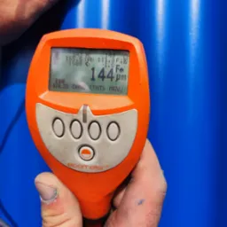 Pomarańczowy miernik grubości powłoki Elcometer w dłoniach, wyświetlający wynik 144μm na tle niebieskiej powierzchni. Widoczne przyciski i menu urządzenia.