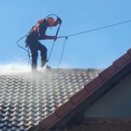 Pracownik w uprzęży i kasku myje dach ciśnieniowo, widoczna para wodna i dachówka.