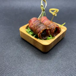 Dwa daktyle zawinięte w bekon, spięte ozdobnymi wykałaczkami, ułożone na rukoli w kwadratowej, bambusowej miseczce na ciemnoszarym tle.