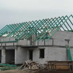 Surowy stan domu z białych bloczków z widoczną konstrukcją dachu z impregnowanego drewna w kolorze zielonym, na pierwszym planie stos drewna i resztki materiałów budowlanych.