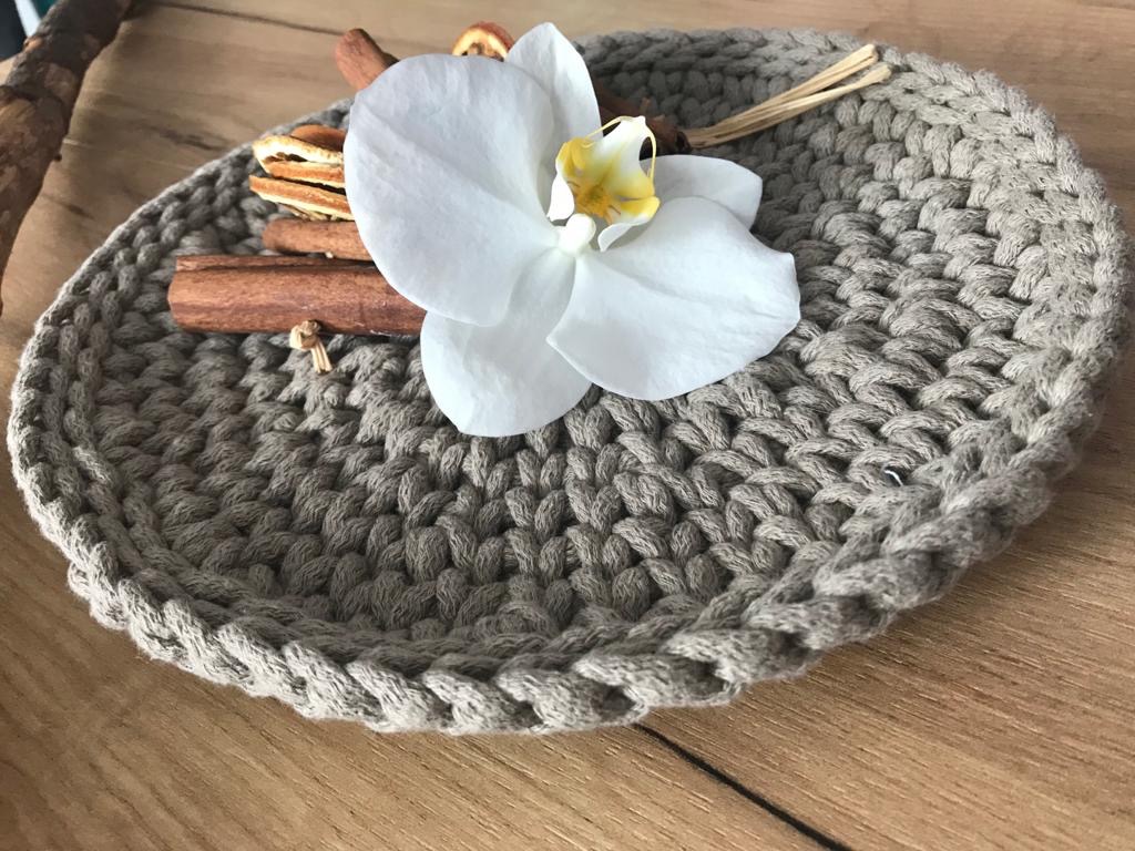 Ręcznie robiona, okrągła, beżowa miseczka z grubego sznurka, udekorowana białą orchideą, laską cynamonu i suszonymi plasterkami pomarańczy, leżąca na drewnianym stole.