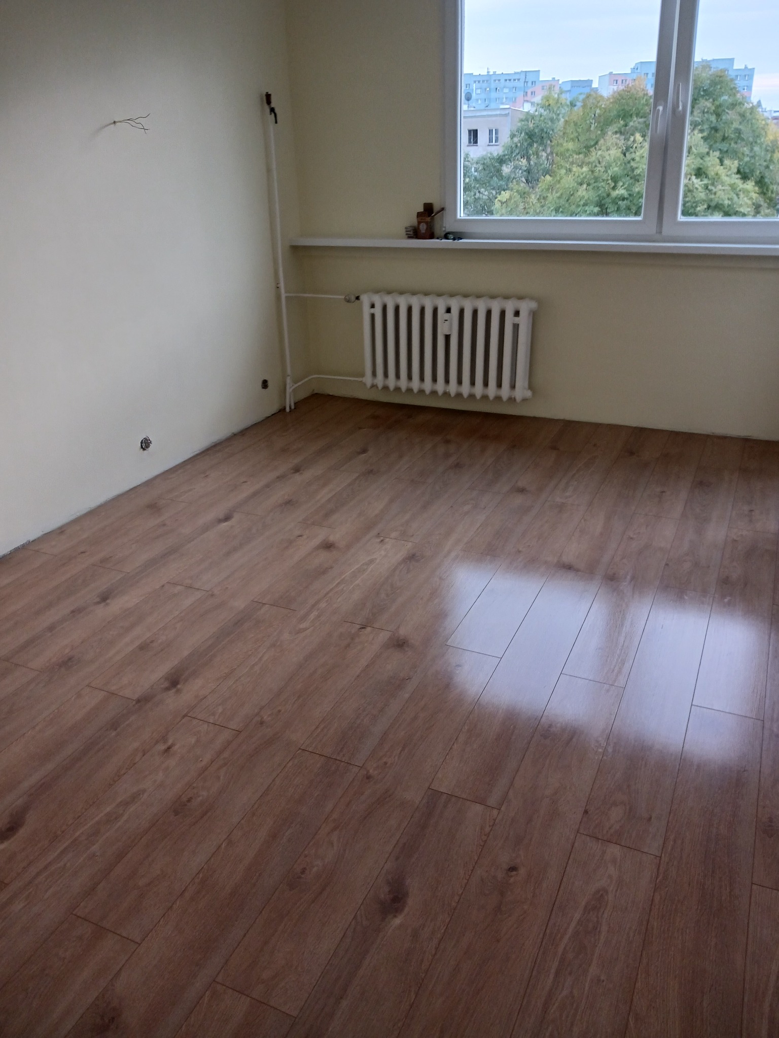 Ułożone panele podłogowe w pokoju z grzejnikiem pod oknem, ściany w trakcie przygotowania do malowania, widoczne wystające kable.