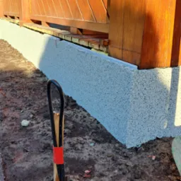 Świeżo nałożona warstwa izolacyjna na fundamencie drewnianego ogrodzenia, widoczny fragment narzędzia z czerwoną taśmą, ziemia wokół.