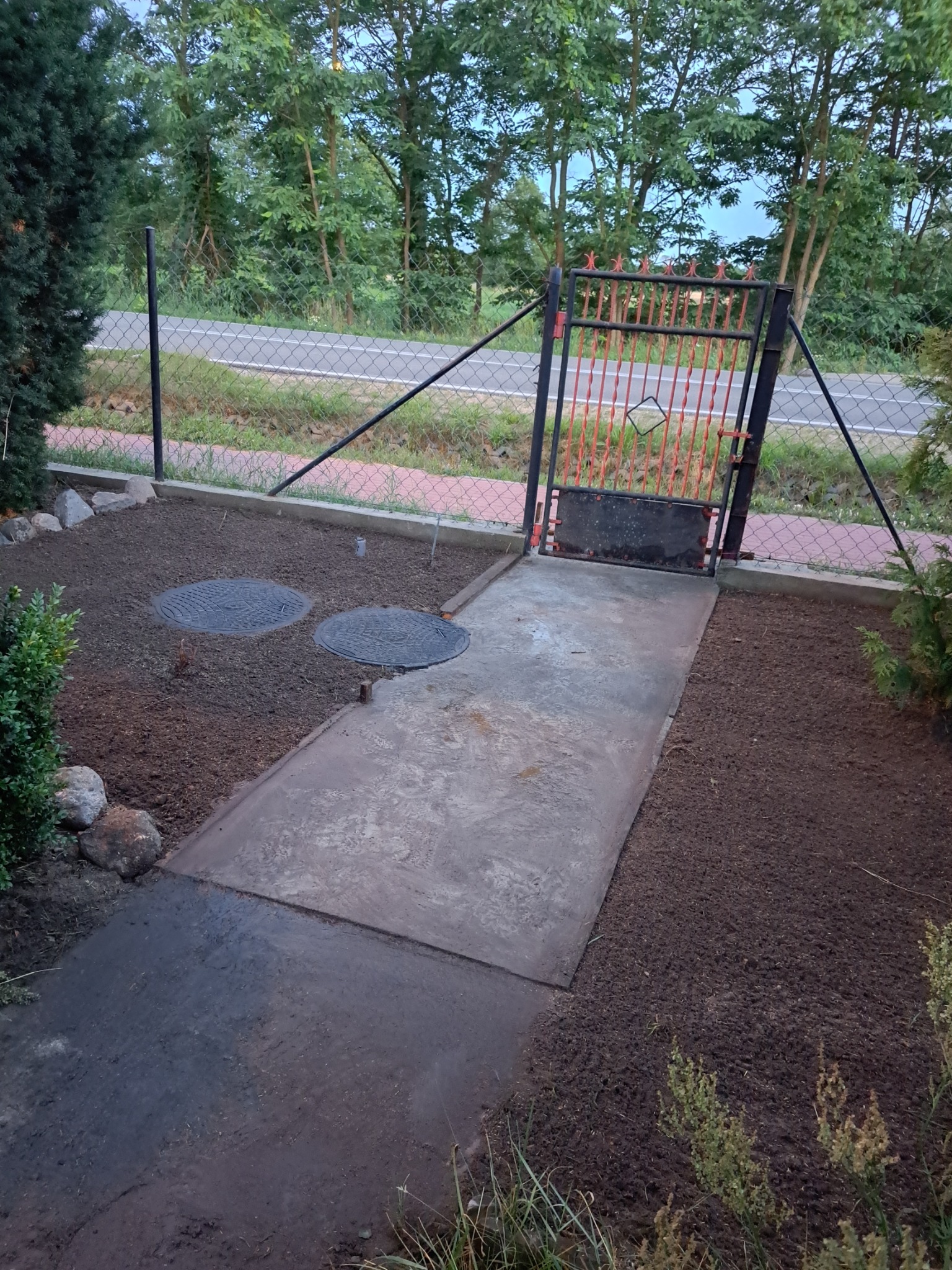 Świeżo wyrównana ziemia w ogrodzie z widocznymi dwoma włazami kanalizacyjnymi, betonowa ścieżka prowadząca do metalowej furtki z pomarańczowymi elementami, w tle siatka ogrodzeniowa i droga.