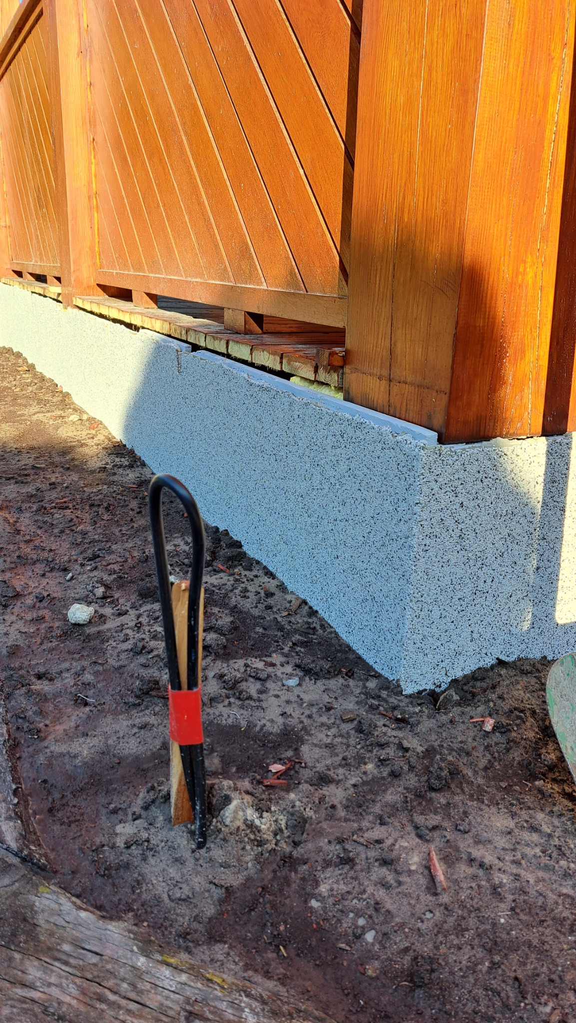 Świeżo nałożona warstwa izolacyjna na fundamencie drewnianego ogrodzenia, widoczny fragment narzędzia z czerwoną taśmą, ziemia wokół.