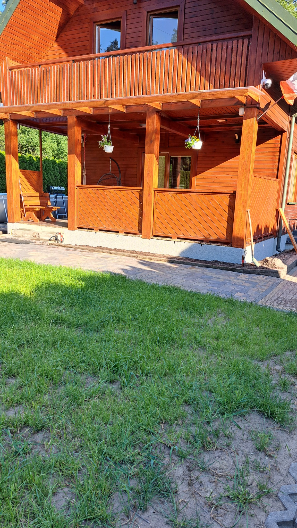 Drewniany dom z werandą, na której wiszą doniczki z kwiatami, widok z trawnika, częściowo ocieniony, na pierwszym planie ślady prac ziemnych.