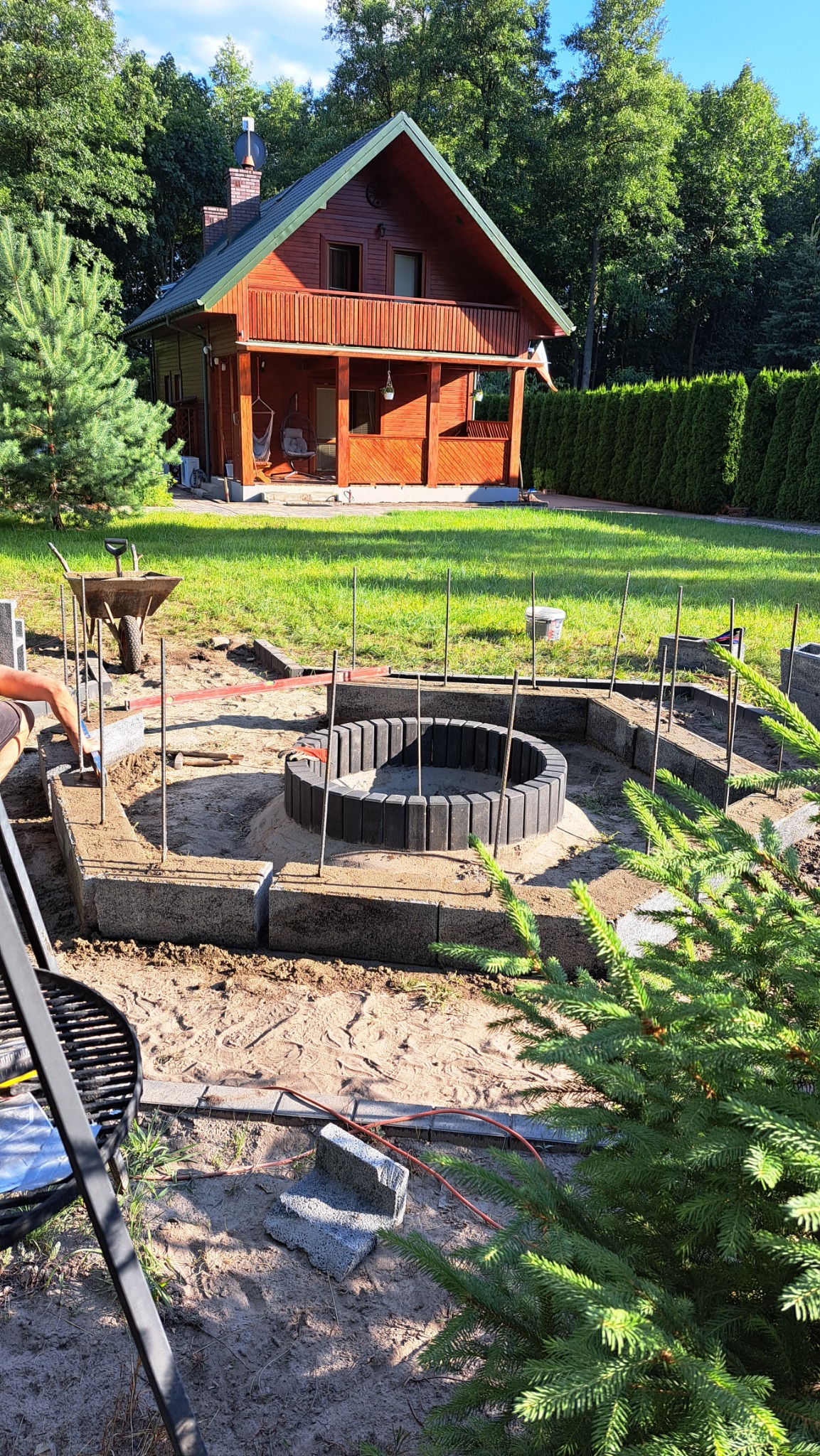 Budowa miejsca na ognisko z betonowych elementów w Kalinowcu, widoczne metalowe pręty stabilizujące konstrukcję, taczka i drewniany dom w tle.