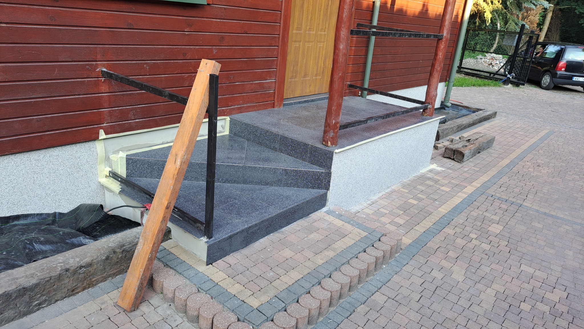 Wykończone schody wejściowe do drewnianego domu z ciemnymi płytkami i szarą elewacją, widoczna drewniana deska oparta o barierkę, kostka brukowa na podjeździe.