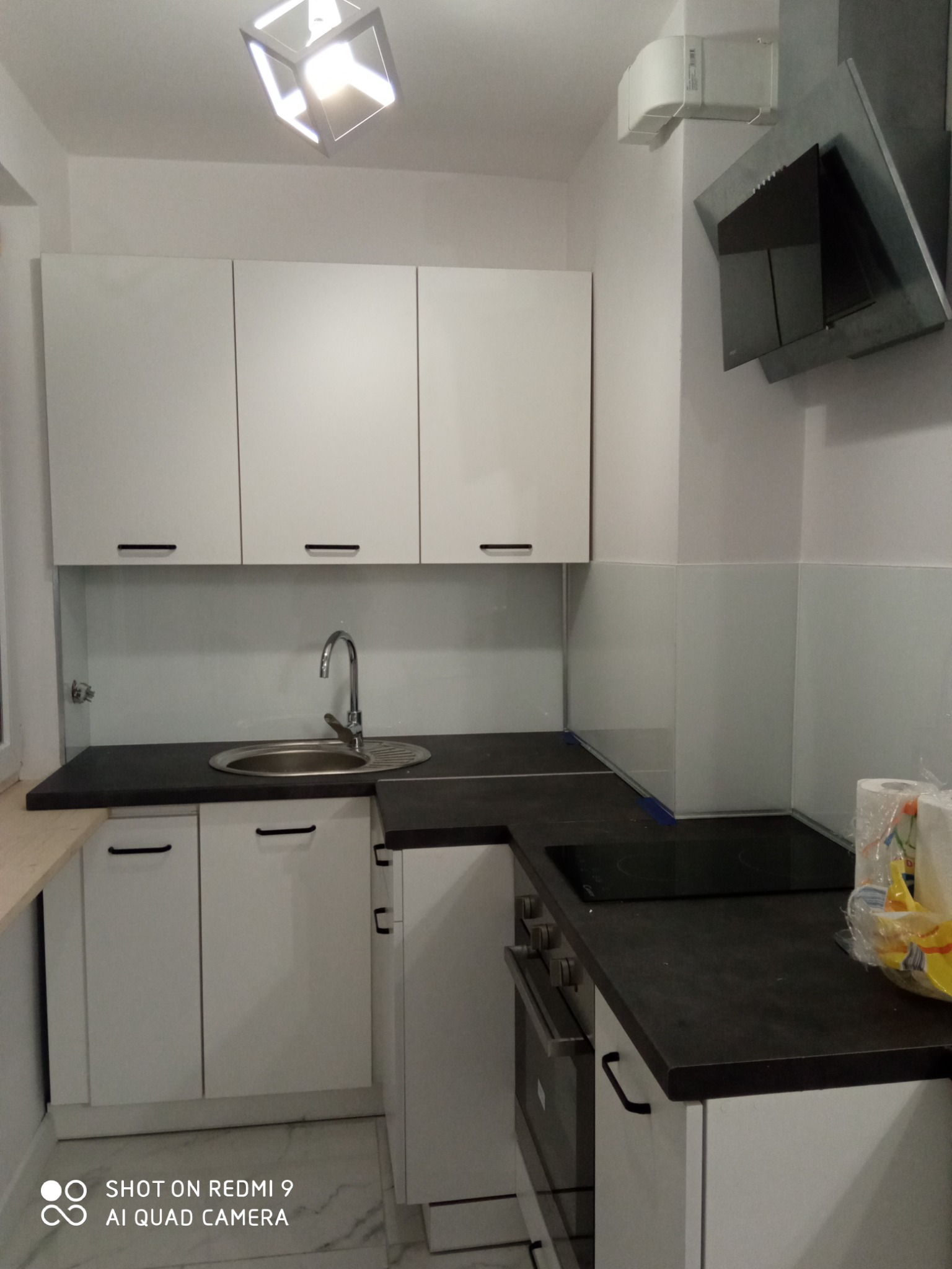 Nowoczesna, minimalistyczna kuchnia z białymi szafkami, czarnym blatem i szklanym panelem nad blatem. Widoczny zlewozmywak ze srebrną baterią, płyta indukcyjna, piekarnik i okap. Górne szafki bez...