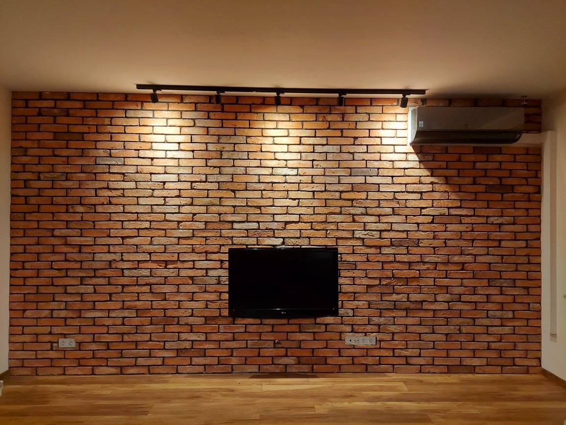 Ściana z cegły klinkierowej w salonie z zamontowanym telewizorem i klimatyzacją, oświetlona reflektorami szynowymi, widoczne gniazdka elektryczne i parkiet na podłodze.