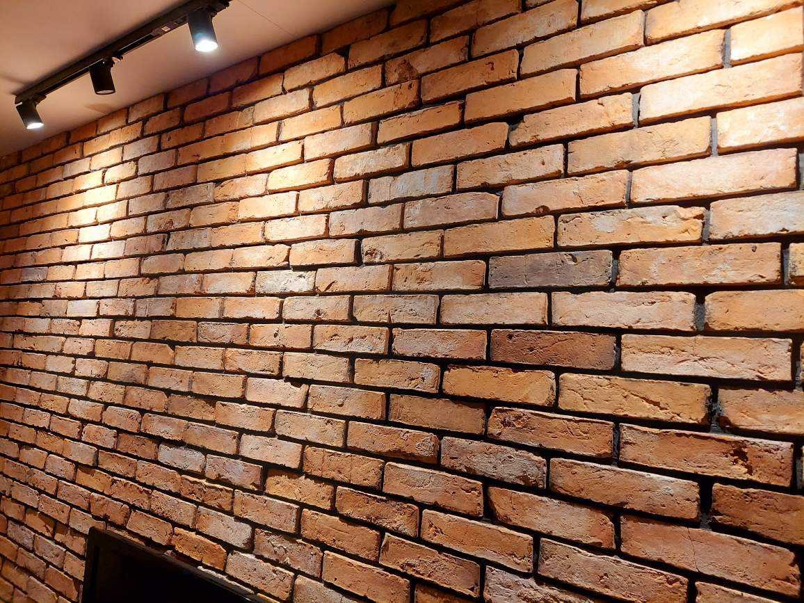 Ściana z cegły klinkierowej w odcieniach pomarańczy i brązu, oświetlona reflektorami szynowymi, widoczny fragment telewizora w dolnym rogu.