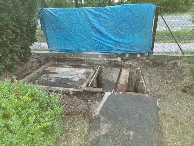 Fundamenty budynku w trakcie prac, zabezpieczone niebieską plandeką, widoczne drewniane elementy konstrukcyjne i świeży beton.