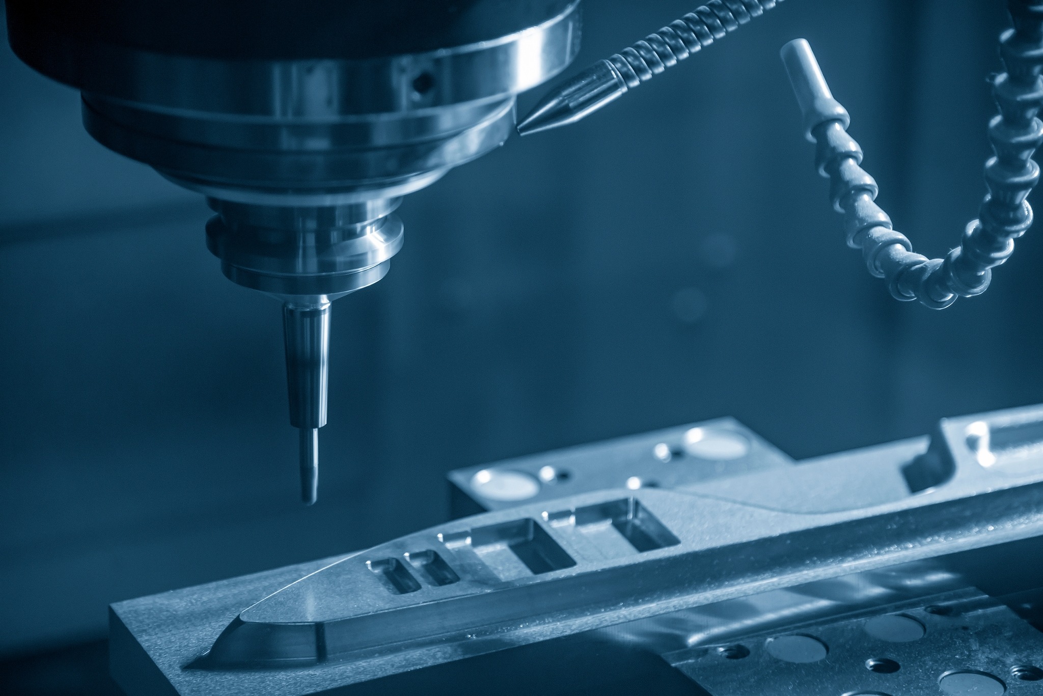 Precyzyjna obróbka CNC metalowego elementu o skomplikowanym kształcie, widoczna głowica frezarki i chłodziwo, całość utrzymana w chłodnej, niebieskiej tonacji.
