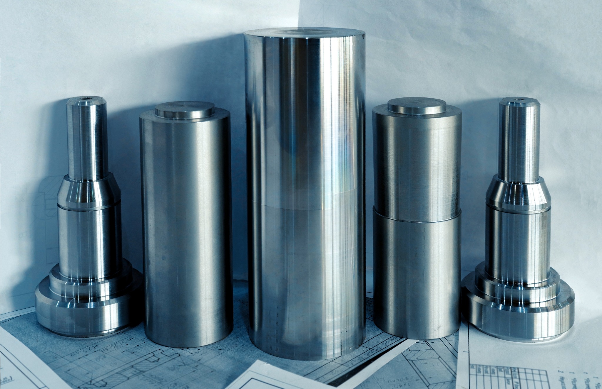 Pięć metalowych cylindrycznych elementów o różnej wysokości, ułożonych na białym tle z widocznymi fragmentami niebieskich rysunków technicznych.