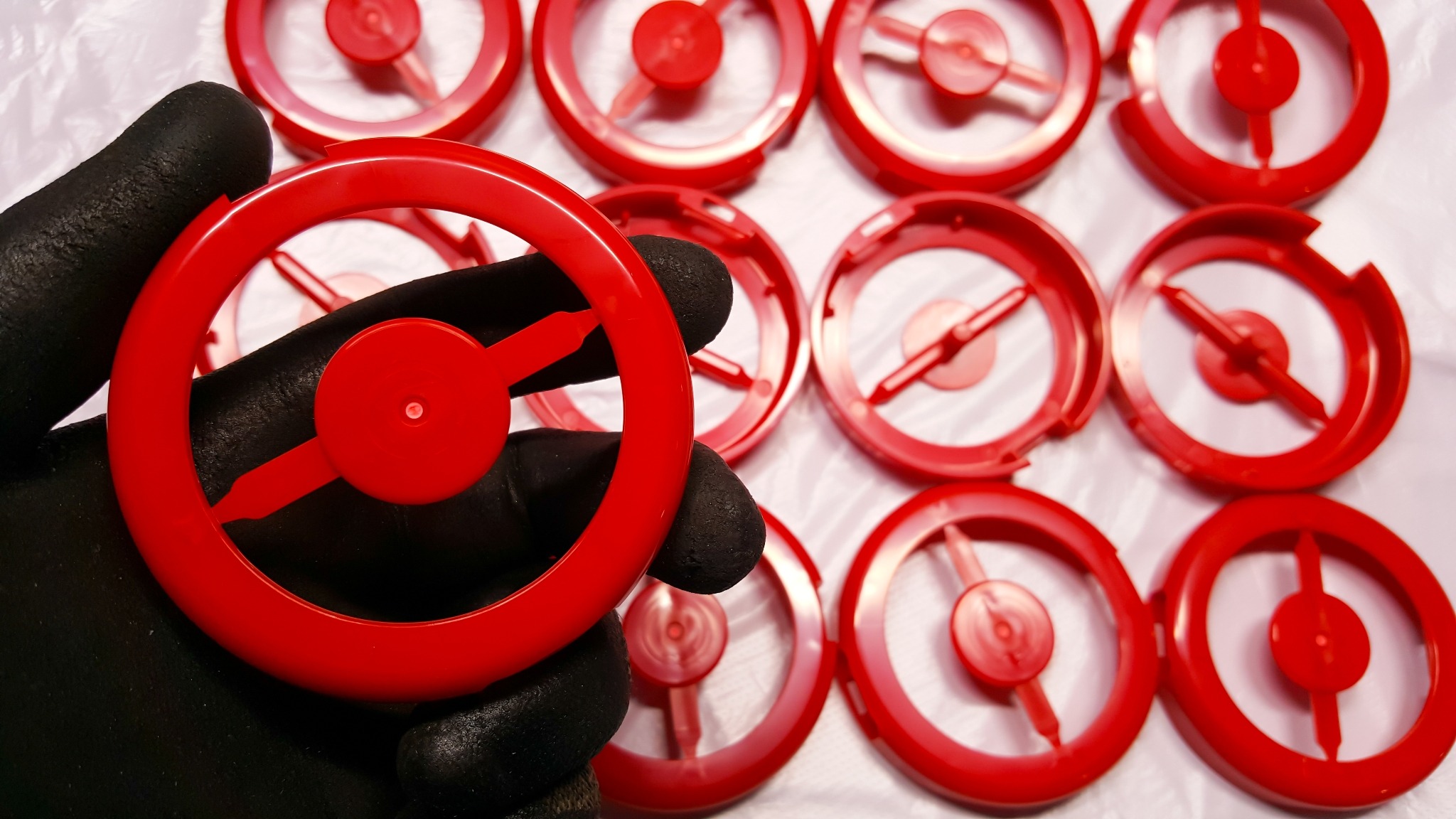 Czerwone, plastikowe elementy z centralnym trzpieniem, trzymane w dłoni w czarnej rękawicy ochronnej, ułożone na białym, lekko pogniecionym podłożu.