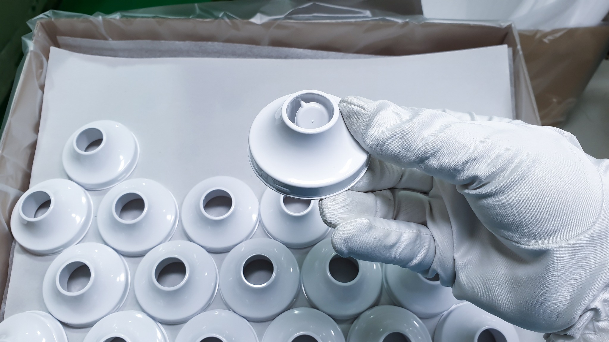 Białe, plastikowe, okrągłe elementy z otworem w środku, układane w kartonowym pudełku wyłożonym papierem, trzymane w dłoni w białej rękawiczce ochronnej.