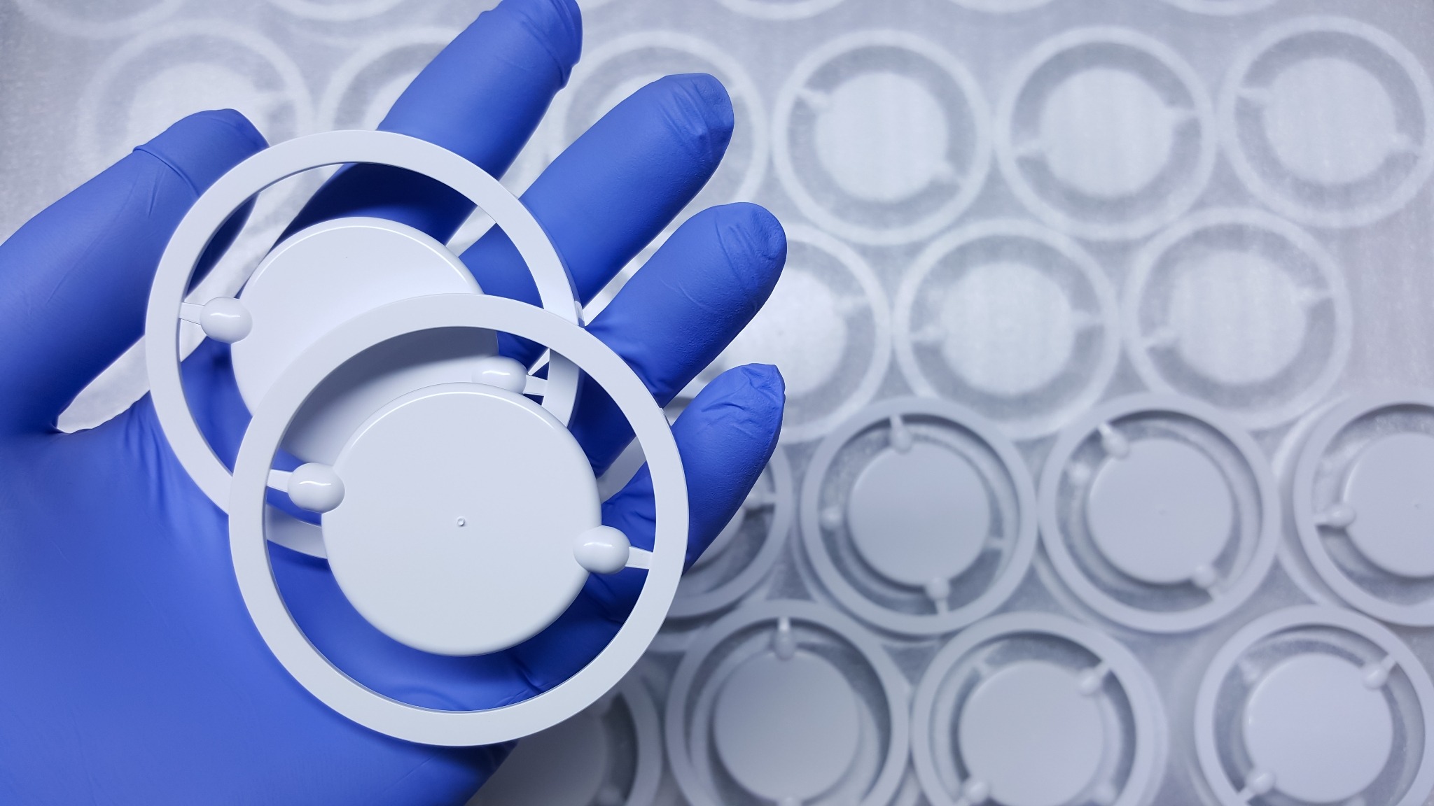 Dłoń w niebieskiej rękawiczce trzyma białe, okrągłe elementy z tworzywa sztucznego na tle wytłoczek z identycznymi elementami.