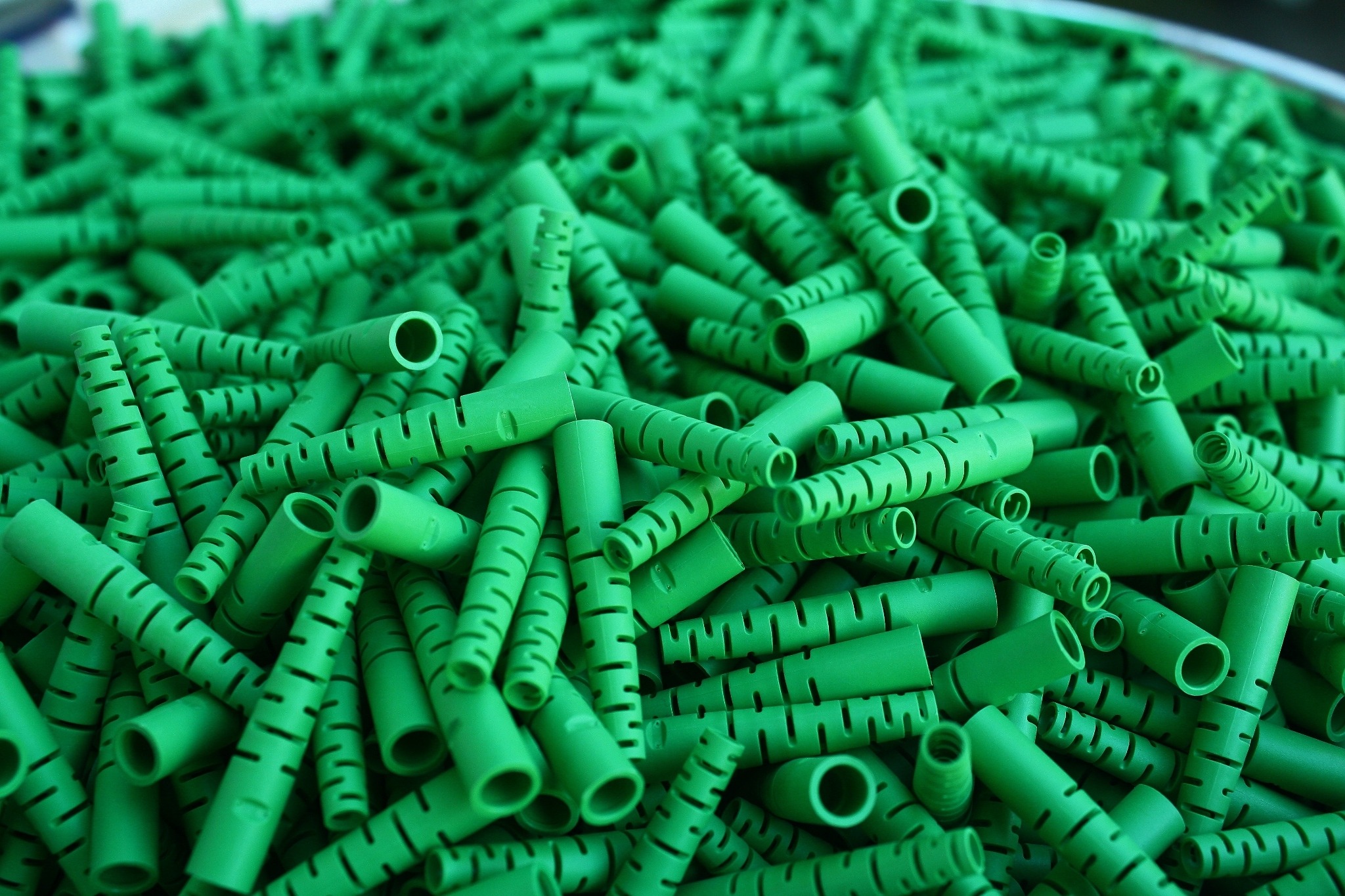Stos zielonych, plastikowych tulejek z nacięciami, ułożonych chaotycznie w pojemniku, widoczne przekroje poprzeczne.