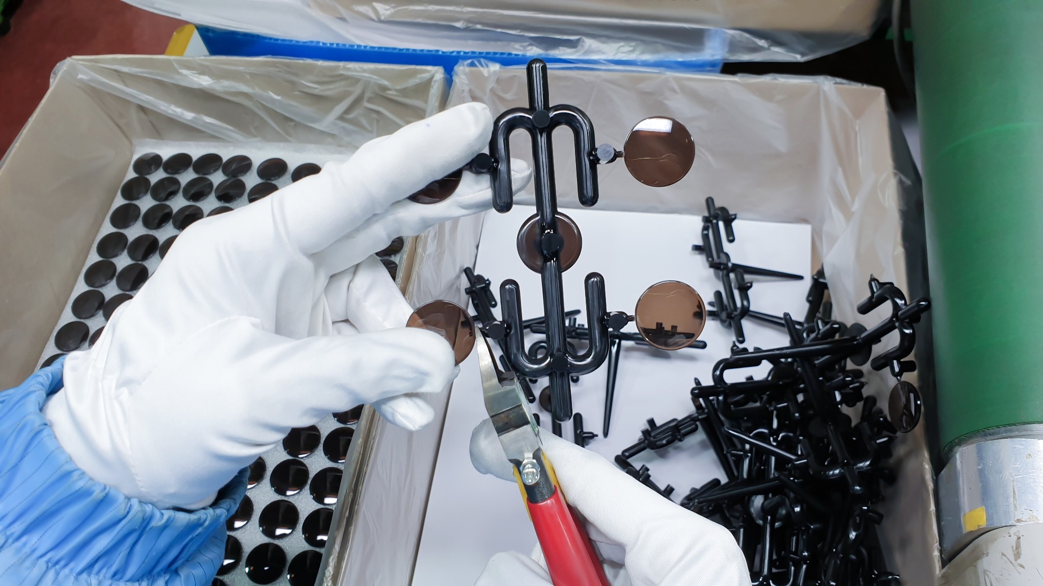 Pracownik w białych rękawiczkach odcina obcęgi od plastikowych ramek okularowych z brązowymi soczewkami, gotowych do zapakowania w opakowania zbiorcze.