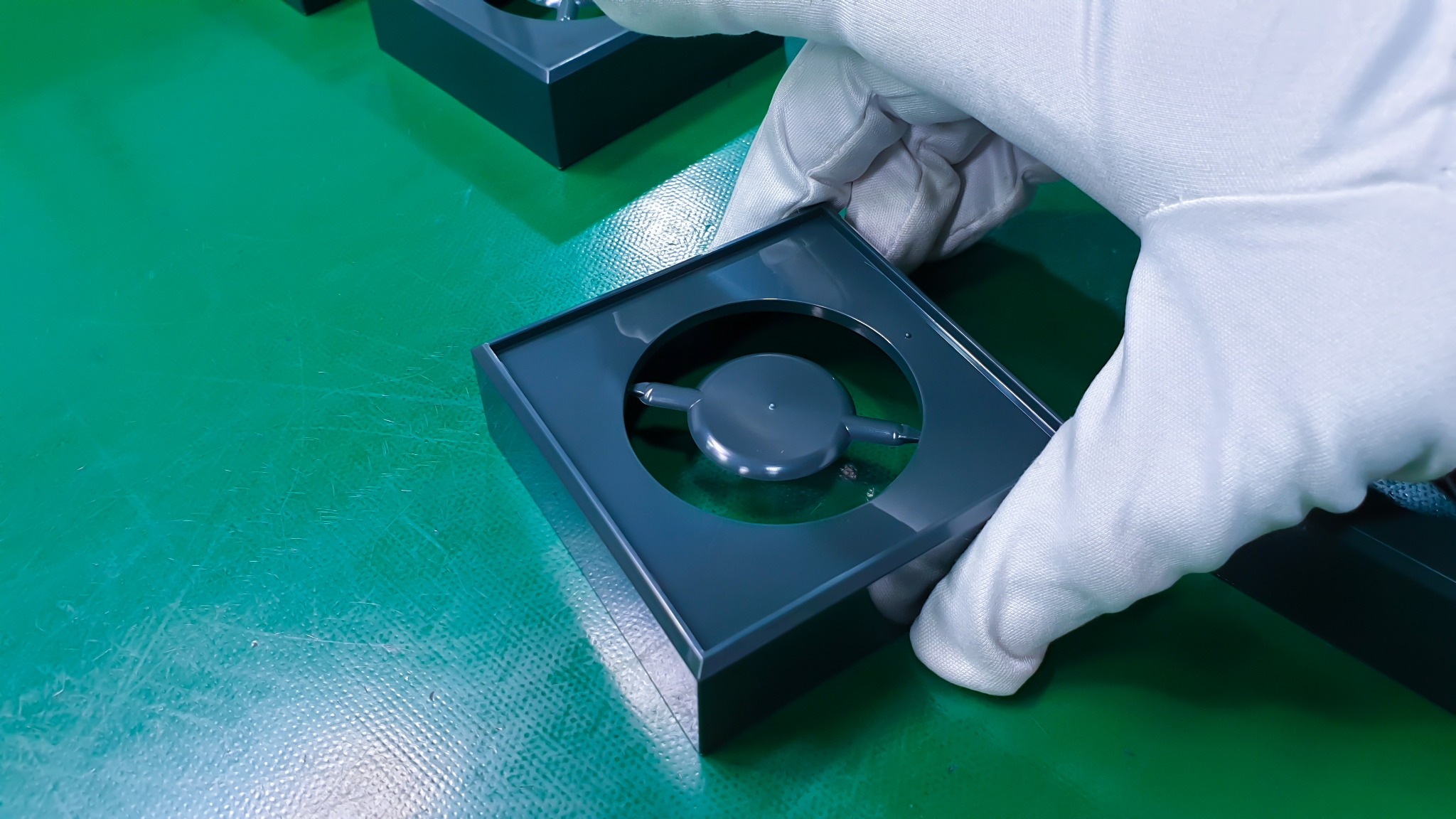Szare pudełko z okrągłym wycięciem na zielonym tle, trzymane w dłoni w białej rękawiczce, prezentujące element wewnątrz.