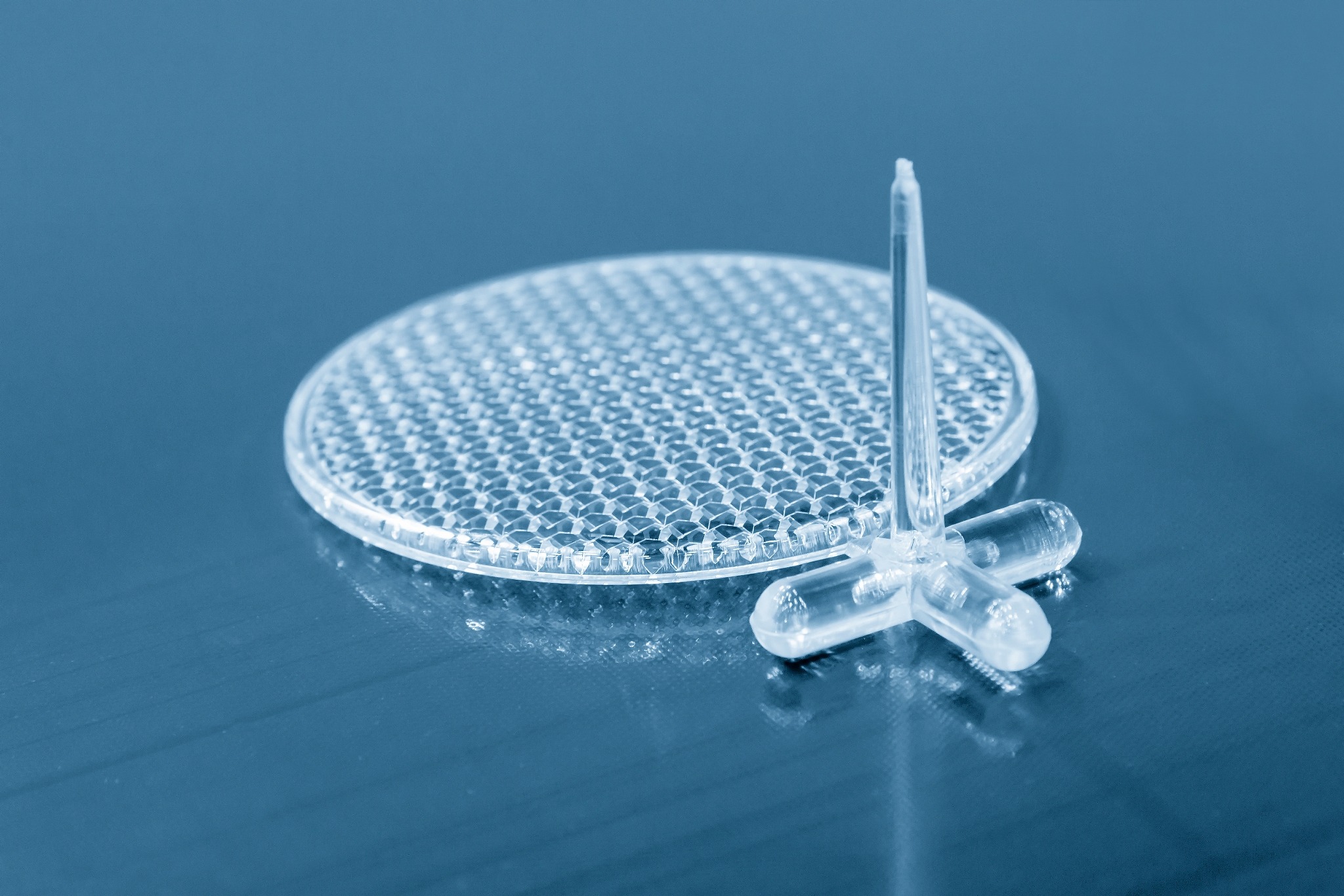 Przezroczysty, okrągły element z tworzywa sztucznego z wzorem plastra miodu i trzema nóżkami, prawdopodobnie element dystansowy lub podkładka, leżący na niebieskim tle.