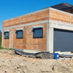 Surowy stan garażu z cegły z zamontowaną szarą bramą garażową segmentową i dwoma oknami z roletami zewnętrznymi, widoczne rusztowanie, teren budowy.