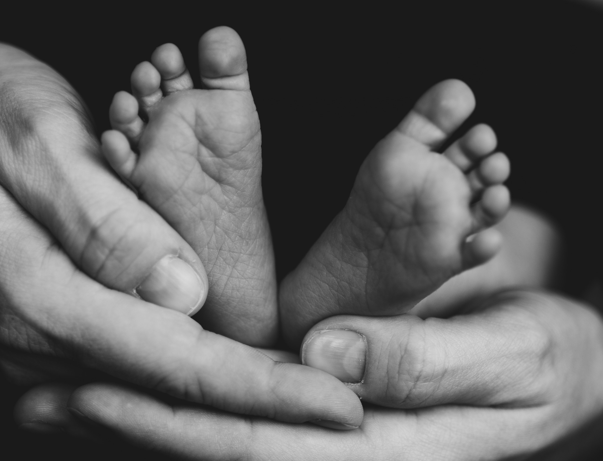 Czarno-białe zbliżenie na małe stopy noworodka trzymane w dłoniach osoby dorosłej, ukazujące delikatne zmarszczki i szczegóły skóry.