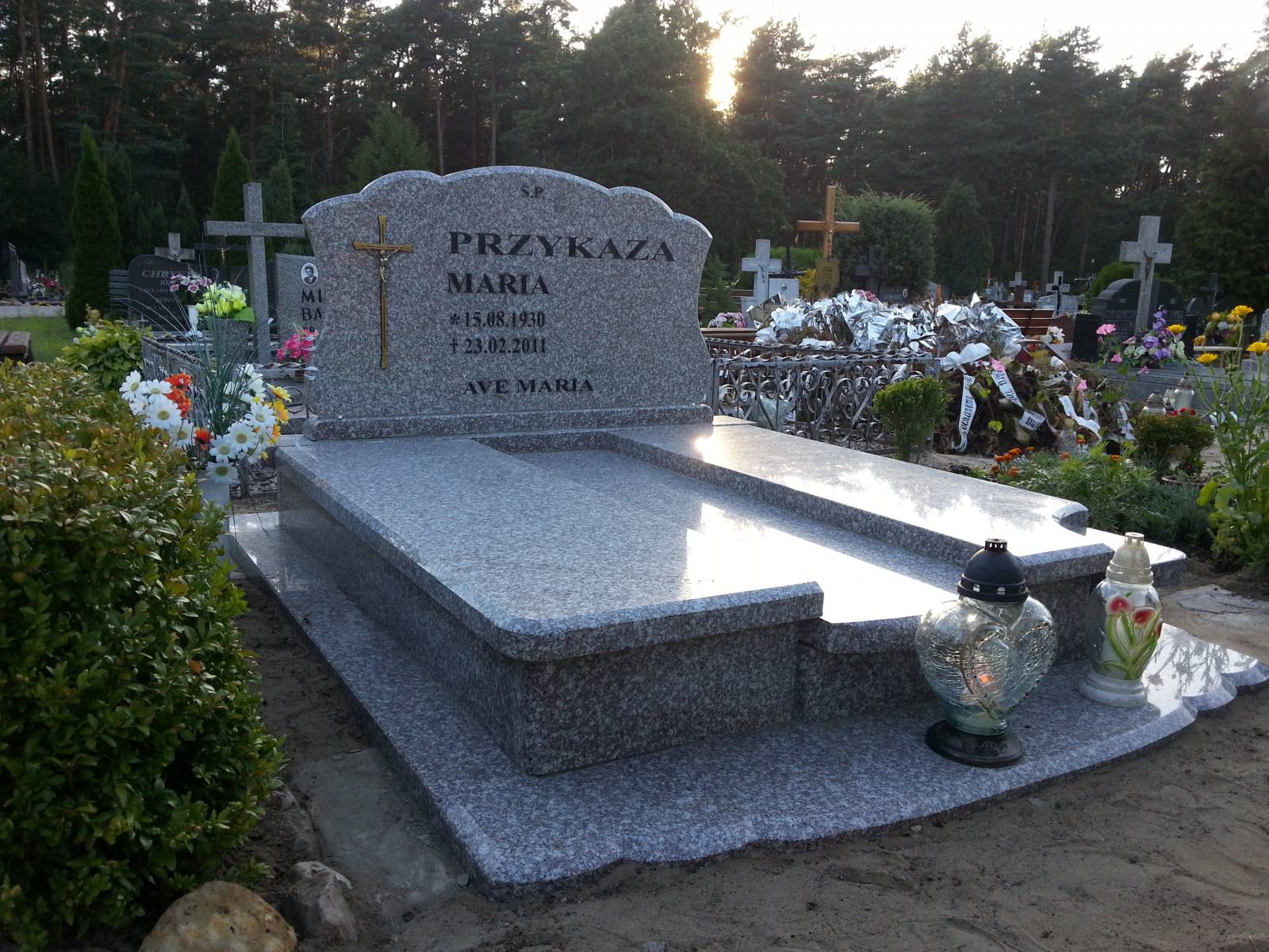 Granitowy nagrobek z wygrawerowanym krzyżem i inskrypcjami na tle cmentarza z licznymi drzewami i nagrobkami, oświetlony popołudniowym słońcem.